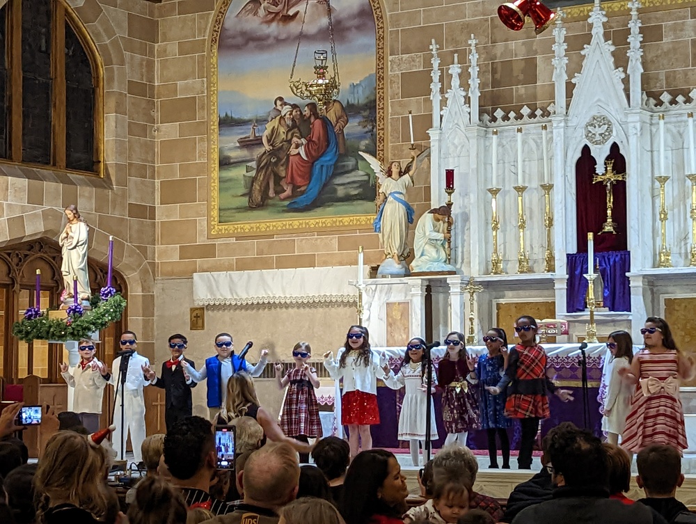 A group of first grade students wearing sunglasses singing in a Catholic Church at the altar