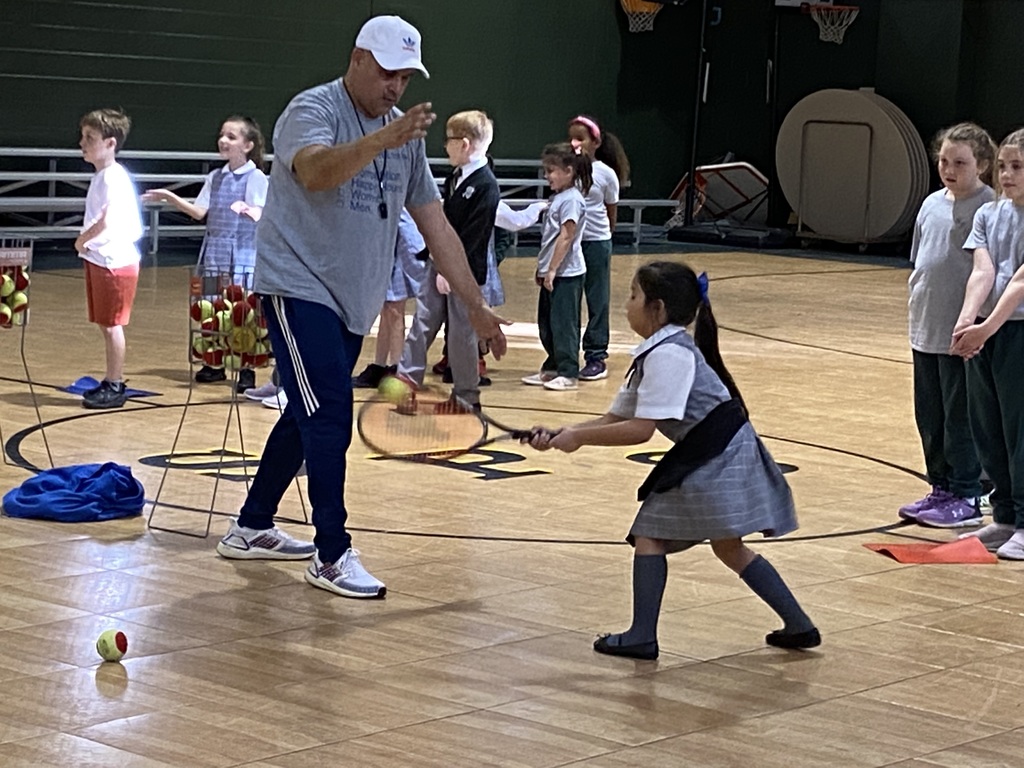 A girl wearing a gray plaid Catholic School Uniform hitting a tennis ball while her coach watches.
